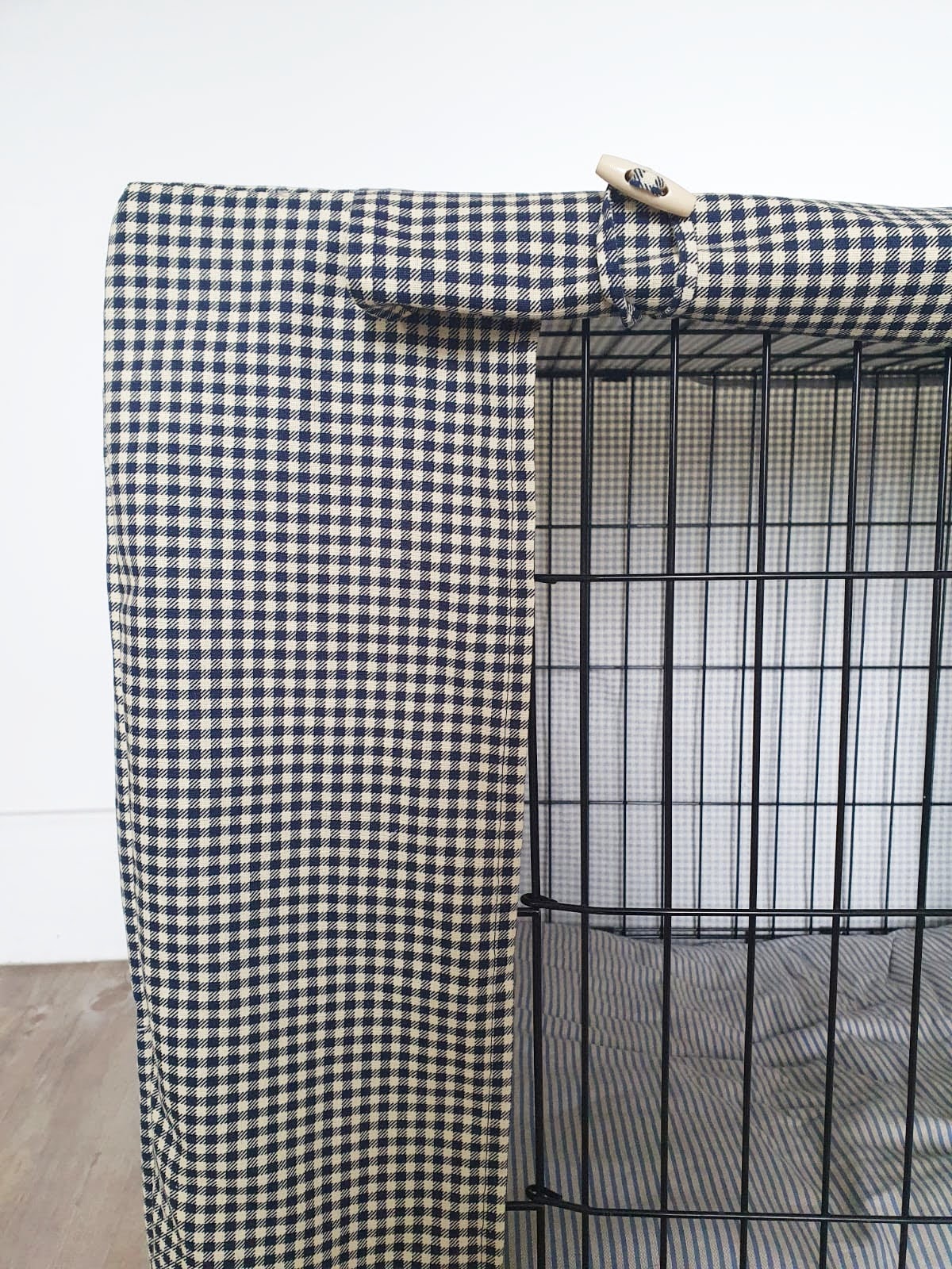 "A black wire dog crate covered with a custom fabric dog crate cover in a navy chequered design. The aesthetic kennel cover features a minimalist design with a roll-up front panel secured by white straps and wooden toggles, providing a cozy and stylish touch to the pet's space."