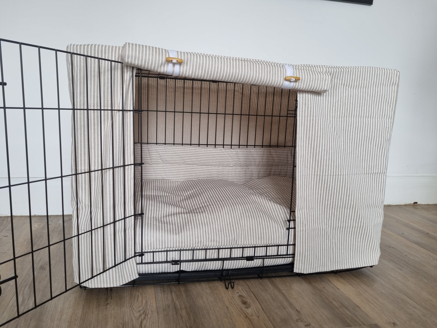 A black wire dog crate covered with a custom fabric dog crate cover in a neutral beige natural striped color. The aesthetic kennel cover features a minimalist design with a roll-up front panel secured by white straps and wooden toggles, providing a cozy and stylish touch to the pet's space.