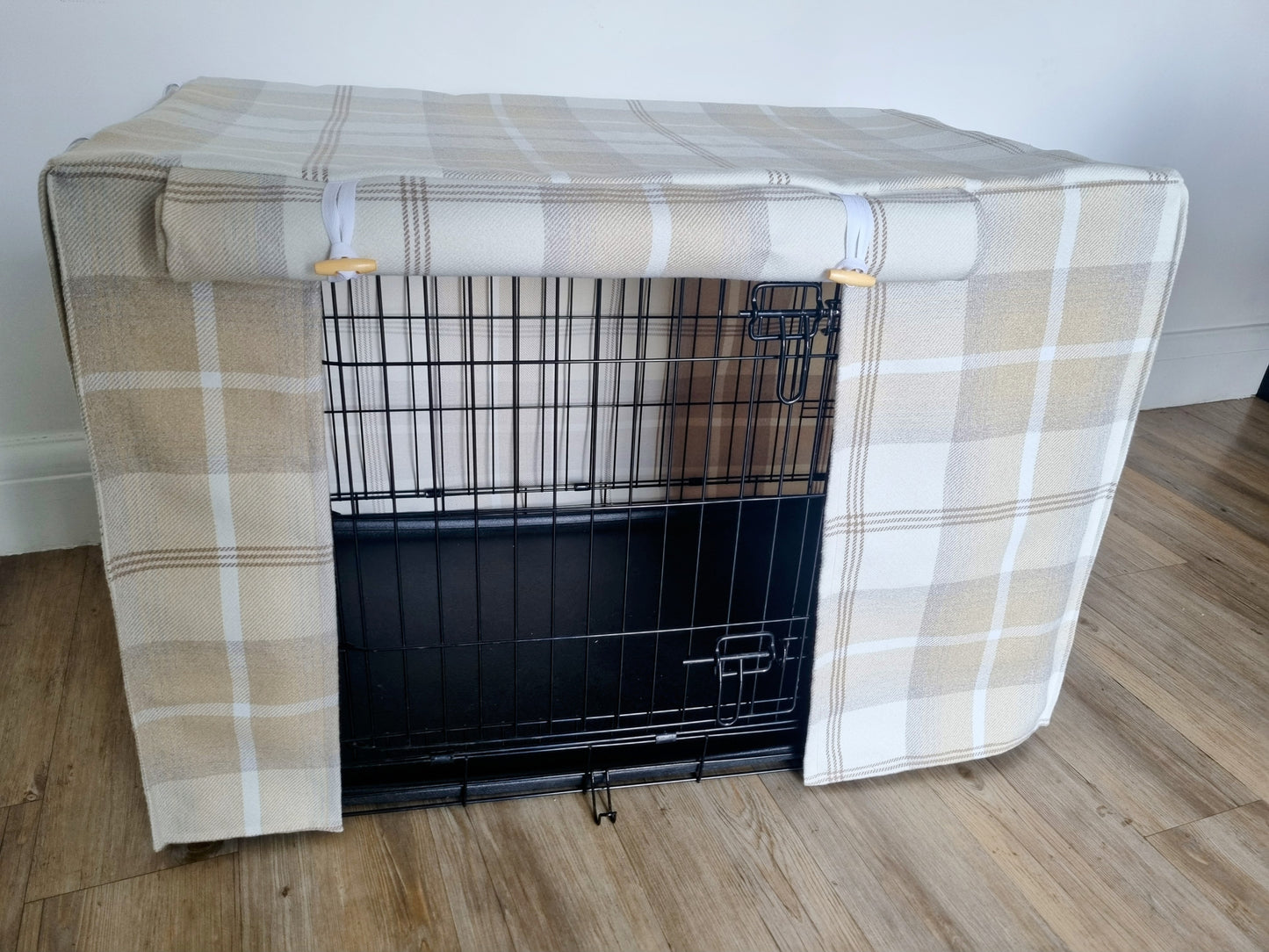 "A black wire dog crate covered with a custom fabric dog crate cover in a neutral cream chequered design. The aesthetic kennel cover features a minimalist design with a roll-up front panel secured by white straps and wooden toggles, providing a cozy and stylish touch to the pet's space."