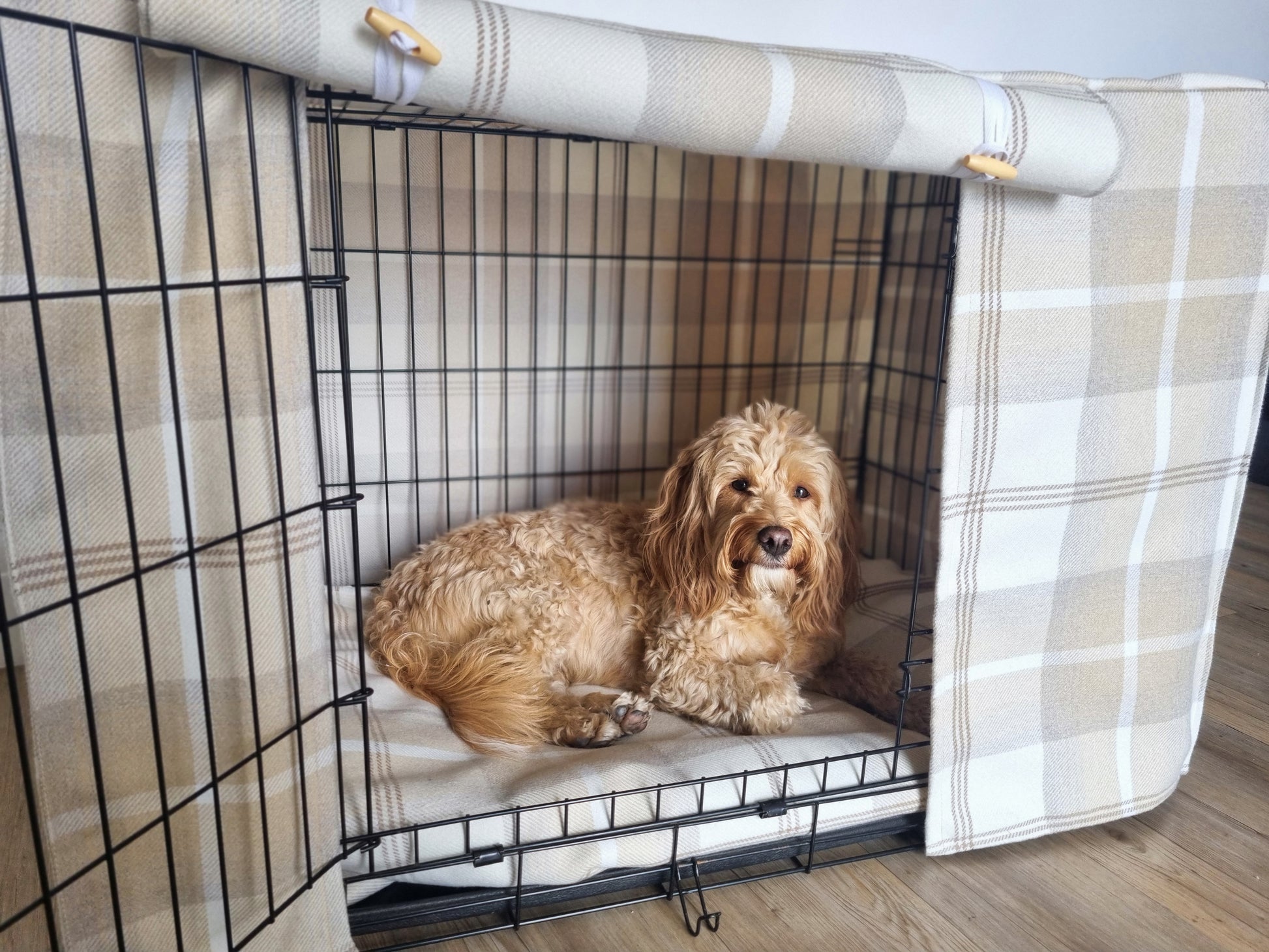 "A black wire dog crate covered with a custom fabric dog crate cover in a neutral cream chequered design. The aesthetic kennel cover features a minimalist design with a roll-up front panel secured by white straps and wooden toggles, providing a cozy and stylish touch to the pet's space."