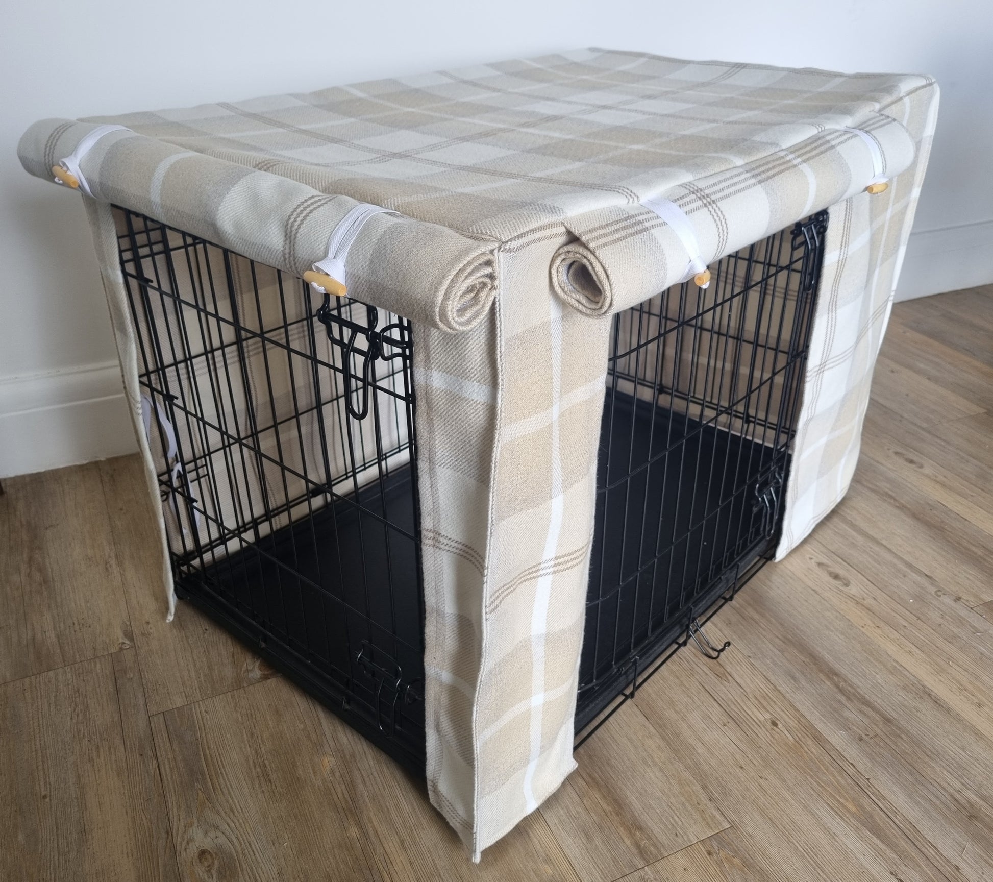 "A black wire dog crate covered with a custom fabric dog crate cover in a neutral cream chequered design. The aesthetic kennel cover features a minimalist design with a roll-up front panel secured by white straps and wooden toggles, providing a cozy and stylish touch to the pet's space."