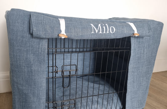 A black wire dog crate covered with a custom fabric dog crate cover in a stylish blue denim-like material. The aesthetic kennel cover features a personalized embroidered name, 'Milo,' on the roll-up front panel, secured with white straps and wooden toggles, creating a cozy and decorative pet space