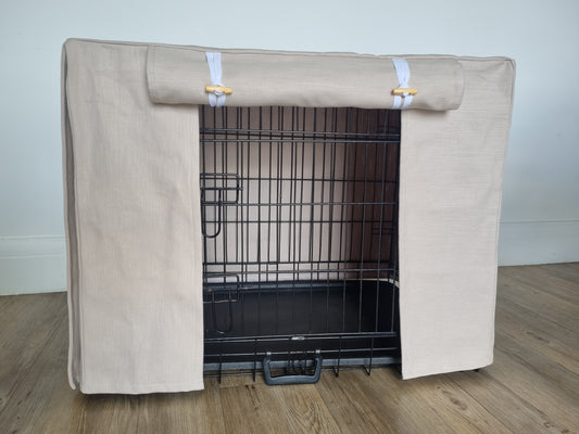 A black wire dog crate covered with a custom fabric dog crate cover in a neutral beige color. The aesthetic kennel cover features a minimalist design with a roll-up front panel secured by white straps and wooden toggles, providing a cozy and stylish touch to the pet's space.