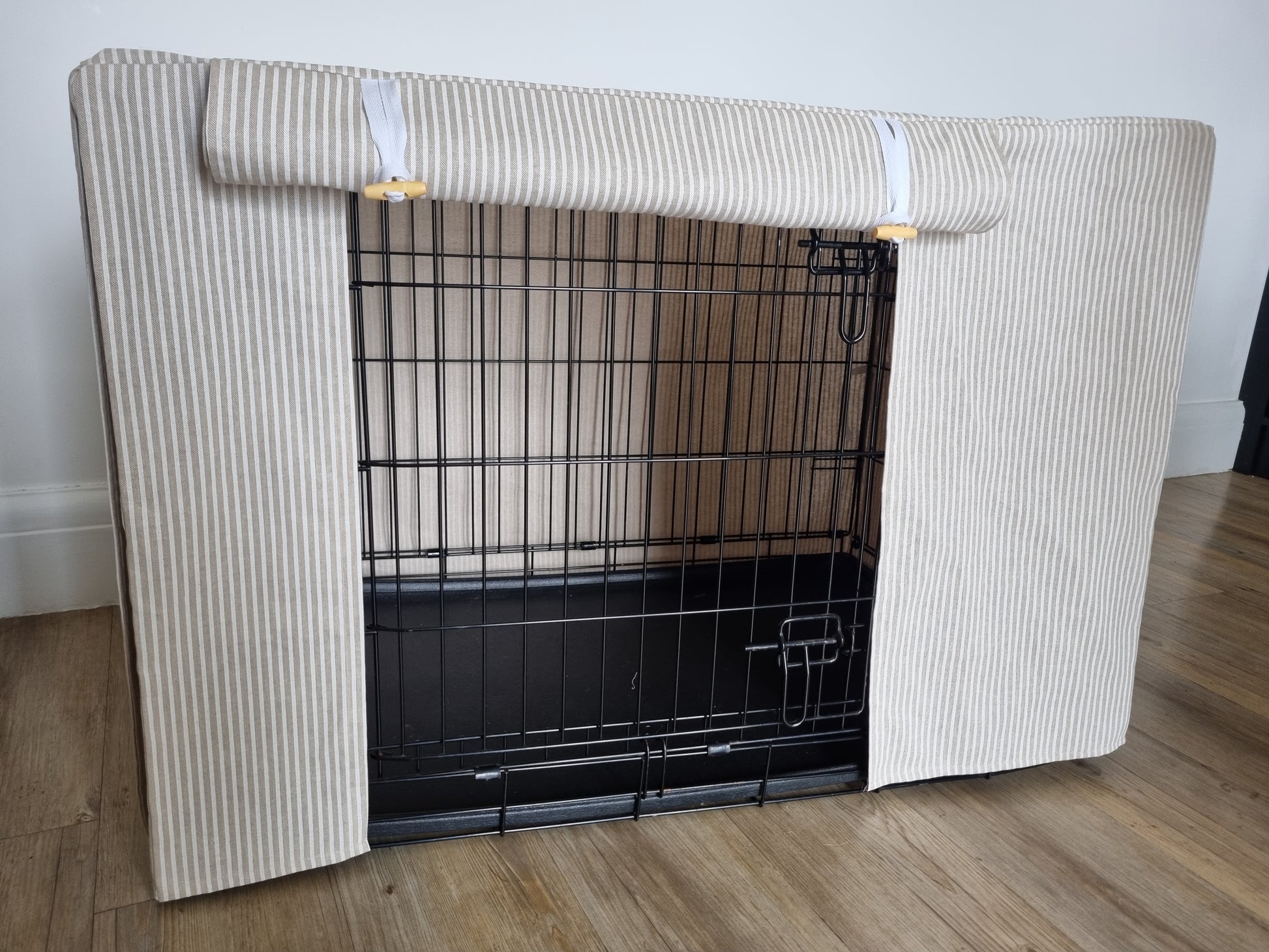 A black wire dog crate covered with a custom fabric dog crate cover in a neutral beige natural striped color. The aesthetic kennel cover features a minimalist design with a roll-up front panel secured by white straps and wooden toggles, providing a cozy and stylish touch to the pet's space.