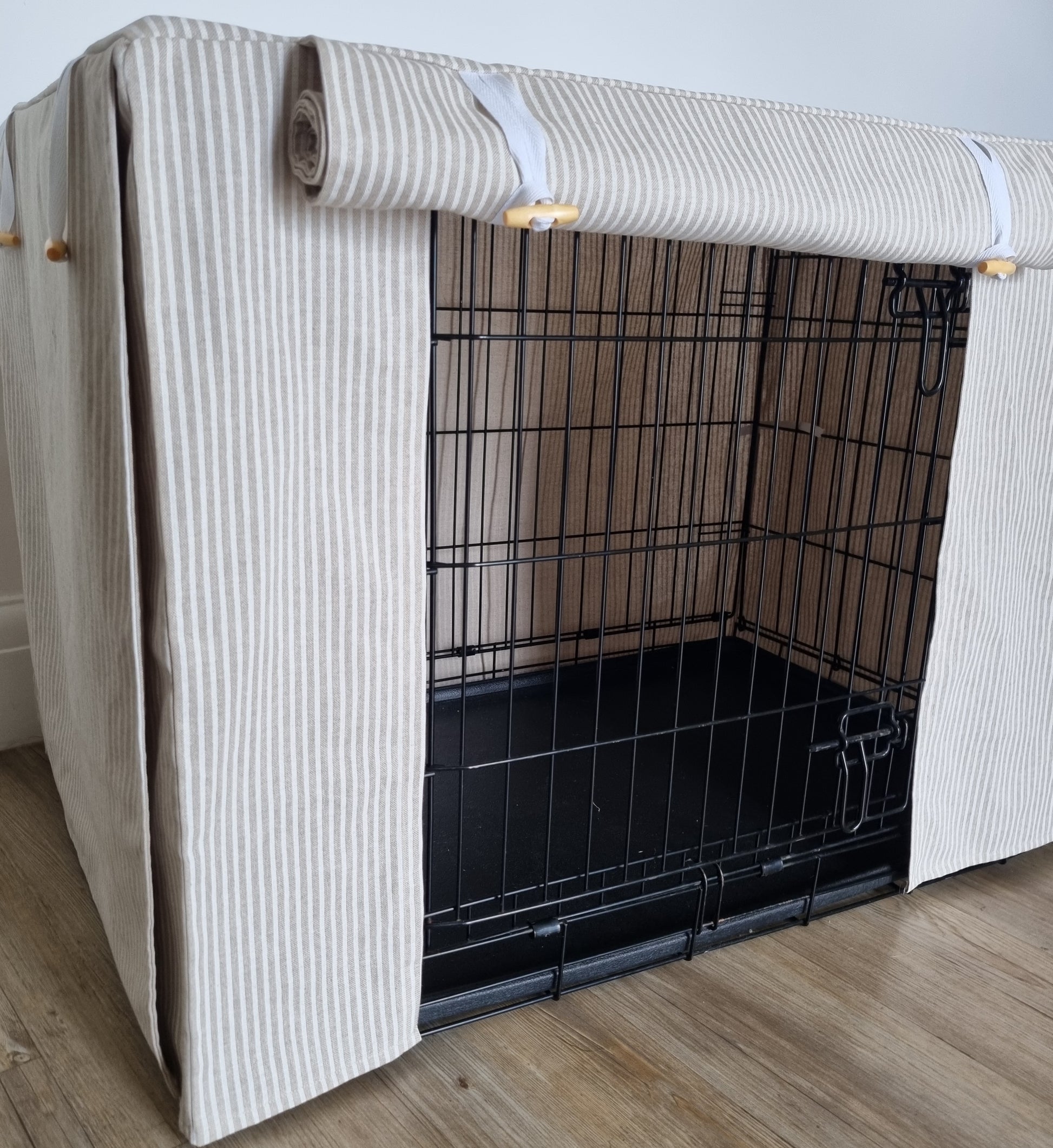 A black wire dog crate covered with a custom fabric dog crate cover in a neutral beige natural striped color. The aesthetic kennel cover features a minimalist design with a roll-up front panel secured by white straps and wooden toggles, providing a cozy and stylish touch to the pet's space.