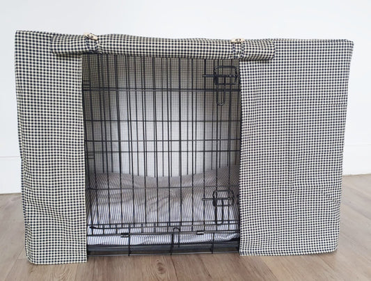 "A black wire dog crate covered with a custom fabric dog crate cover in a navy chequered design. The aesthetic kennel cover features a minimalist design with a roll-up front panel secured by white straps and wooden toggles, providing a cozy and stylish touch to the pet's space."