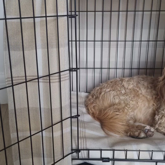 "A black wire dog crate covered with a custom fabric dog crate cover in a neutral cream chequered design. The aesthetic kennel cover features a minimalist design with a roll-up front panel secured by white straps and wooden toggles, providing a cozy and stylish touch to the pet's space."