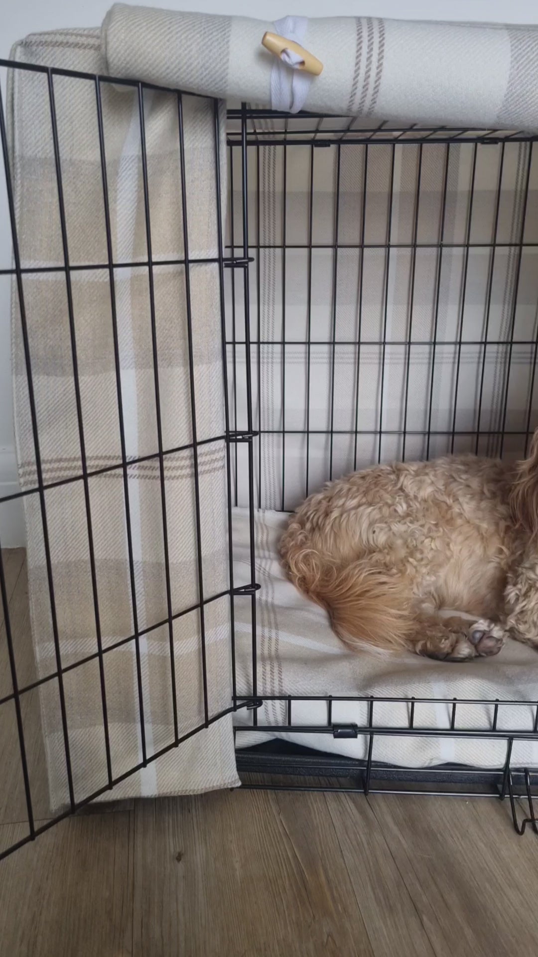 "A black wire dog crate covered with a custom fabric dog crate cover in a neutral cream chequered design. The aesthetic kennel cover features a minimalist design with a roll-up front panel secured by white straps and wooden toggles, providing a cozy and stylish touch to the pet's space."